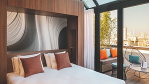 Una habitación de hotel moderna con una cama grande, vistas a la ciudad y un balcón con mobiliario exterior.