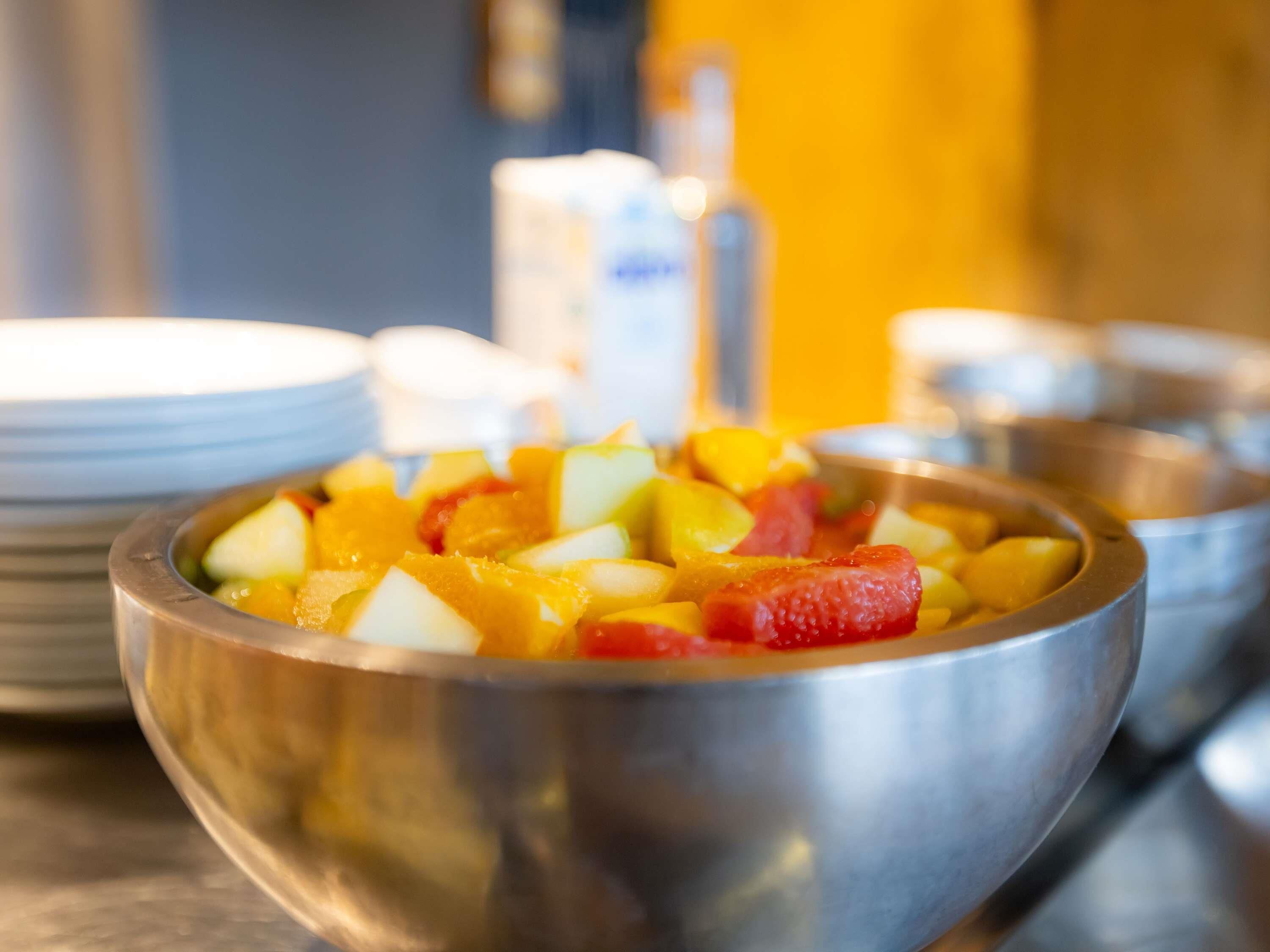 A metal bowl filled with diced fruits, including apples and mangoes, on a table with stacked plates and a blurred background.