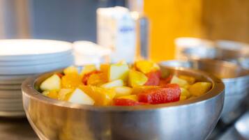 A metal bowl filled with diced fruits, including apples and mangoes, on a table with stacked plates and a blurred background.