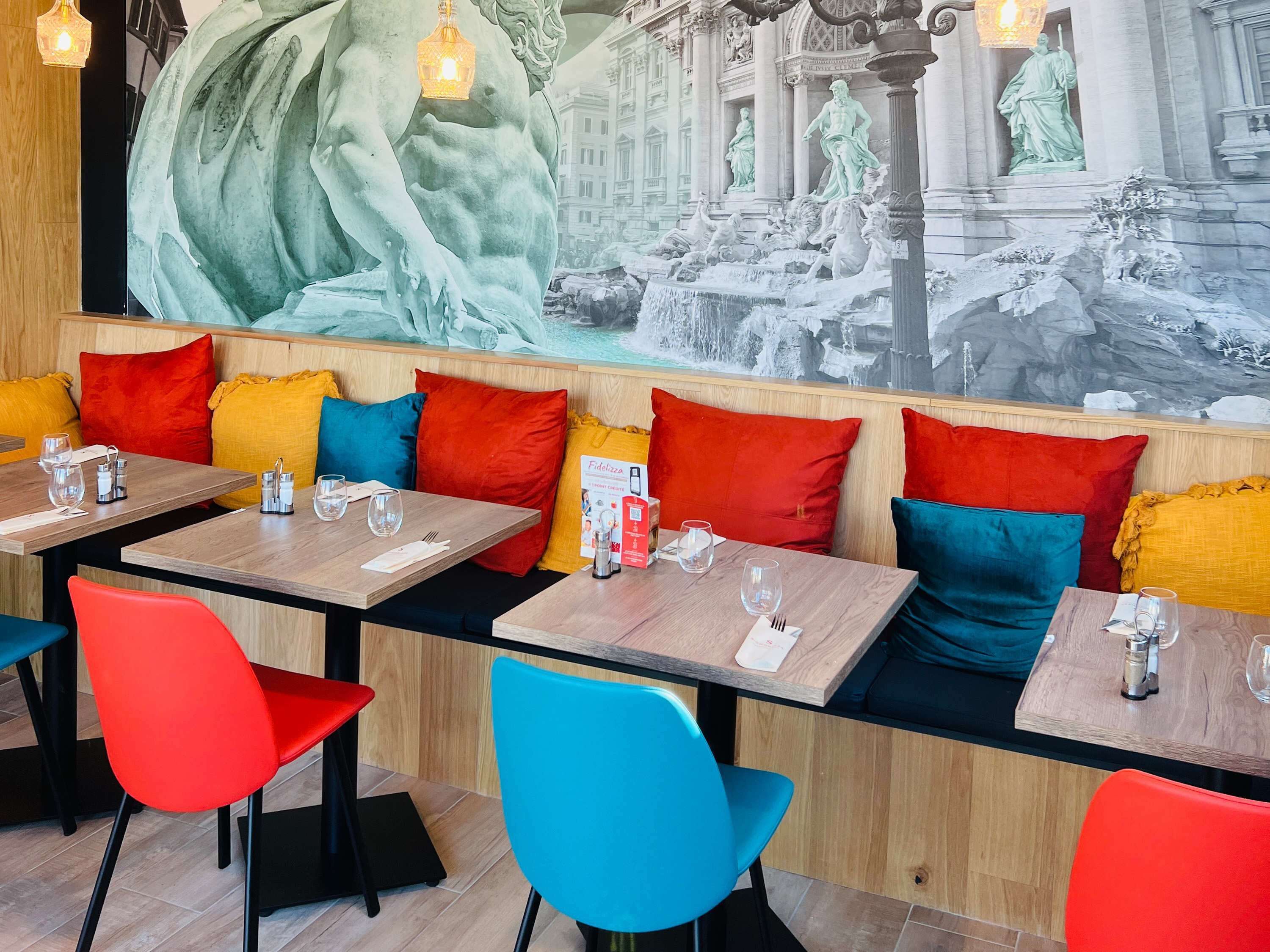 Un restaurante con mesas de madera, sillas coloridas y un gran mural de una fuente y estatuas en la pared.