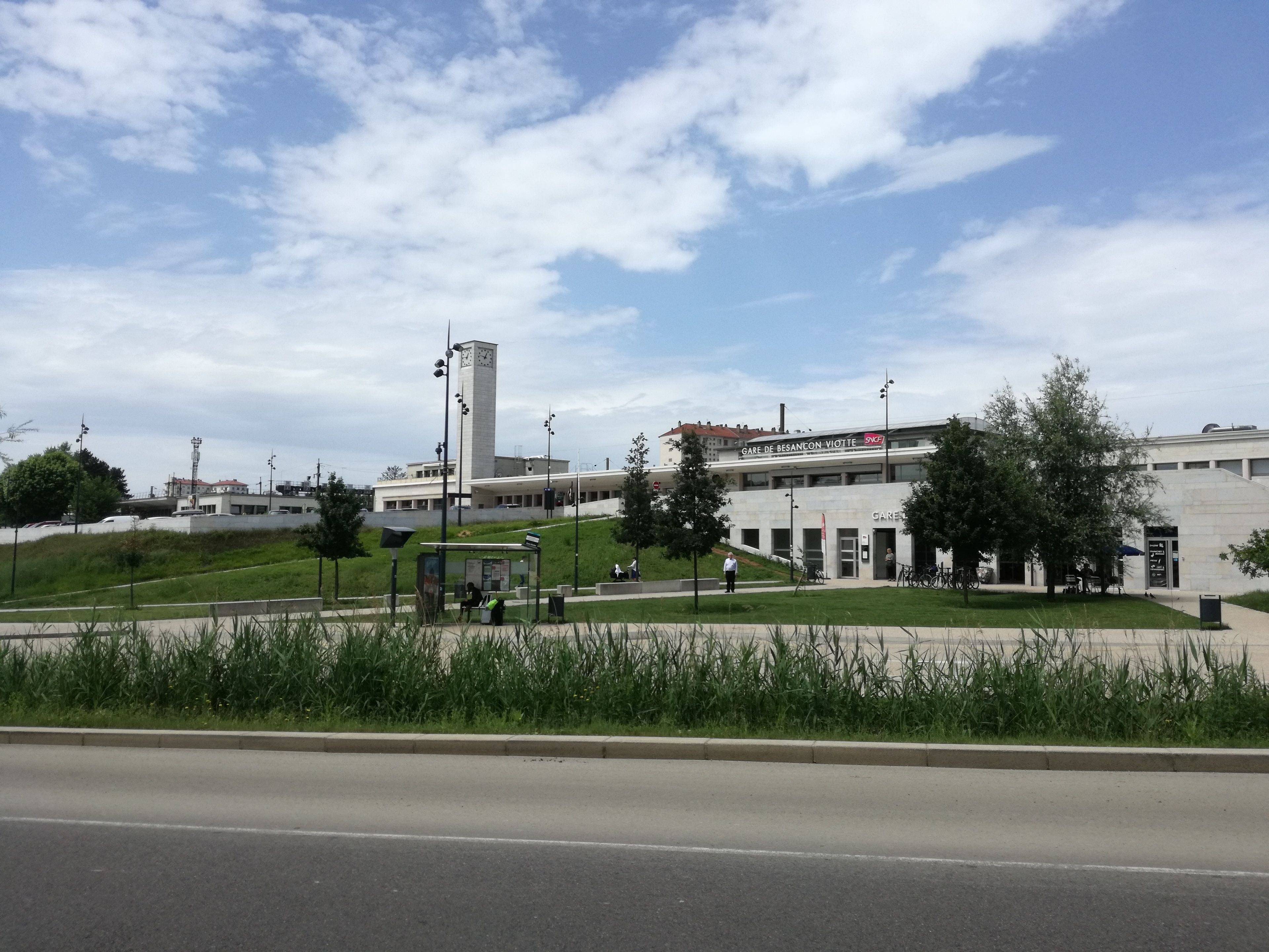 Un edificio con una torre del reloj, un cartel que dice "GARE DE BESANCON VIOTTE" y una zona ajardinada con bancos.