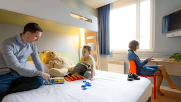 Quarto de hotel com cama, escrivaninha, um urso de pelúcia grande e uma criança brincando com blocos.