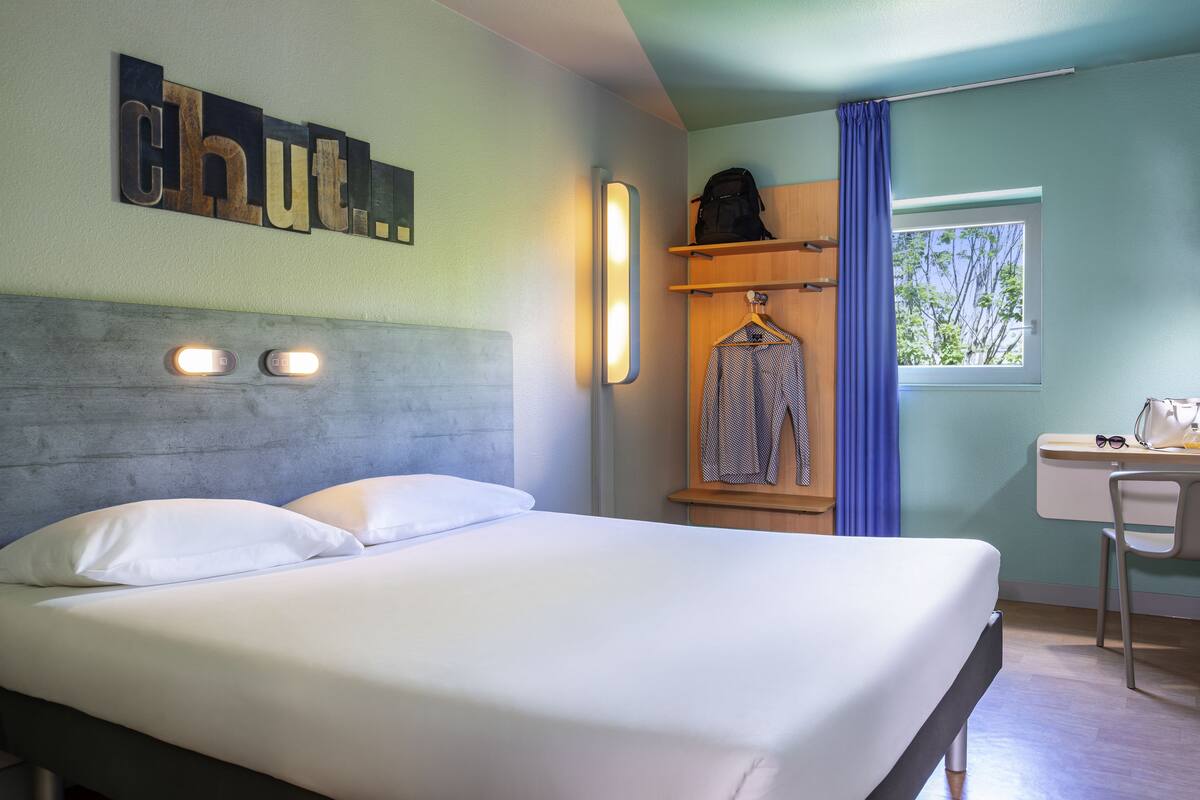 A neatly made bed with white linens, a wooden headboard, and a wall-mounted light fixture.