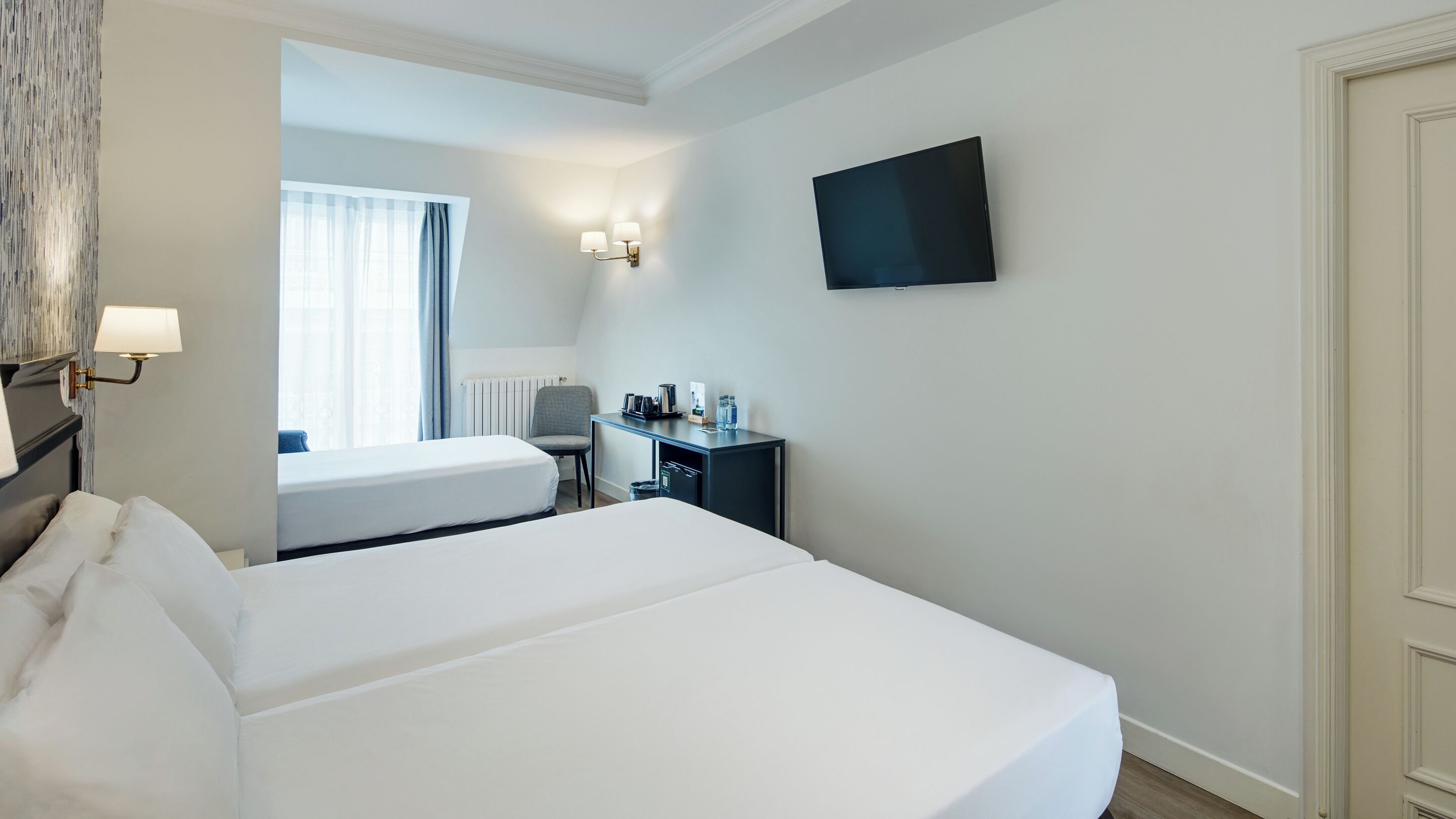 A hotel room with a bed, a desk with a chair, a television, and a window with curtains.