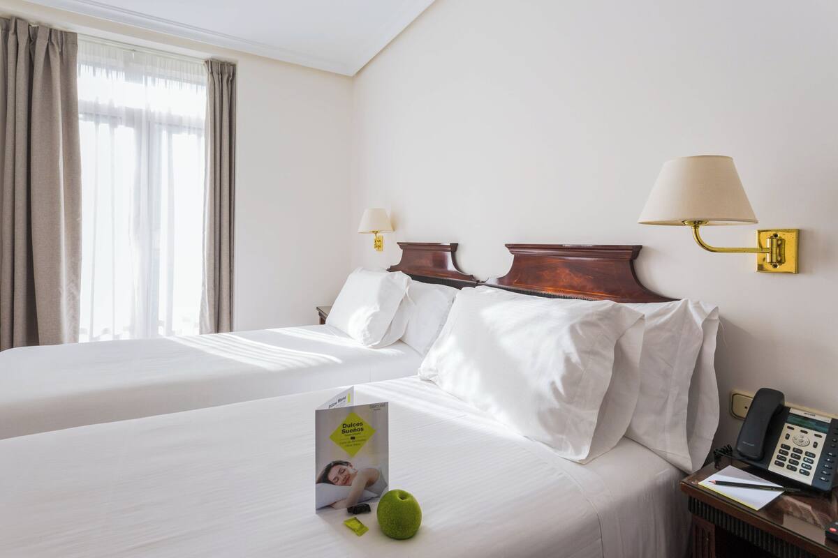 A hotel room with two beds, a bedside table with a lamp, a phone, and a brochure on the bed.