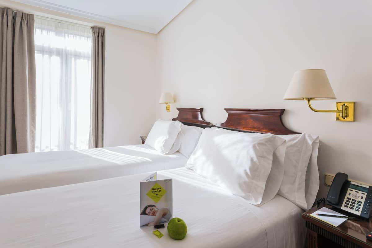 A hotel room with two beds, a bedside table with a lamp, a phone, and a brochure on the bed.