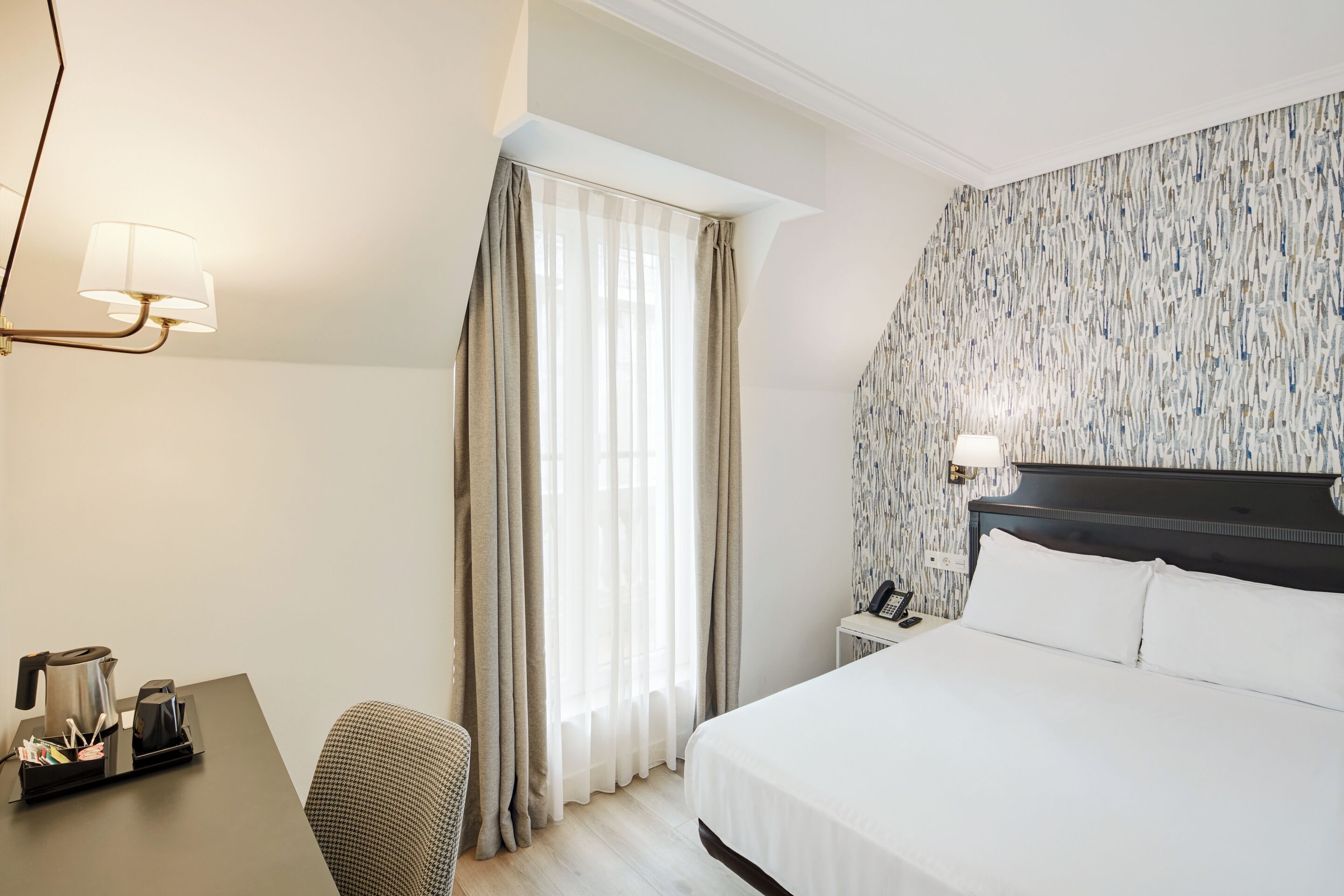 A hotel room with a bed, a desk with a kettle, a chair, and a window with curtains.
