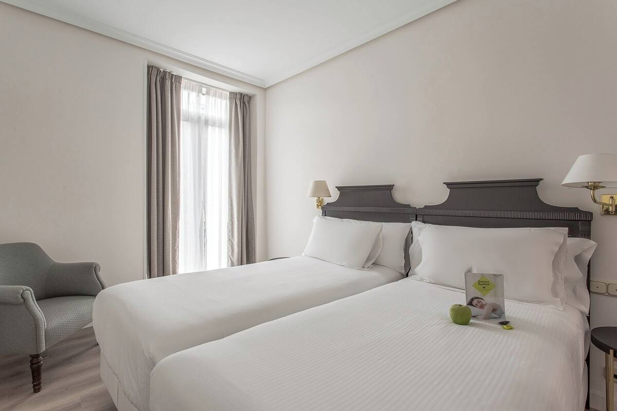 A hotel room with two beds, a chair, a nightstand, and a window with curtains.
