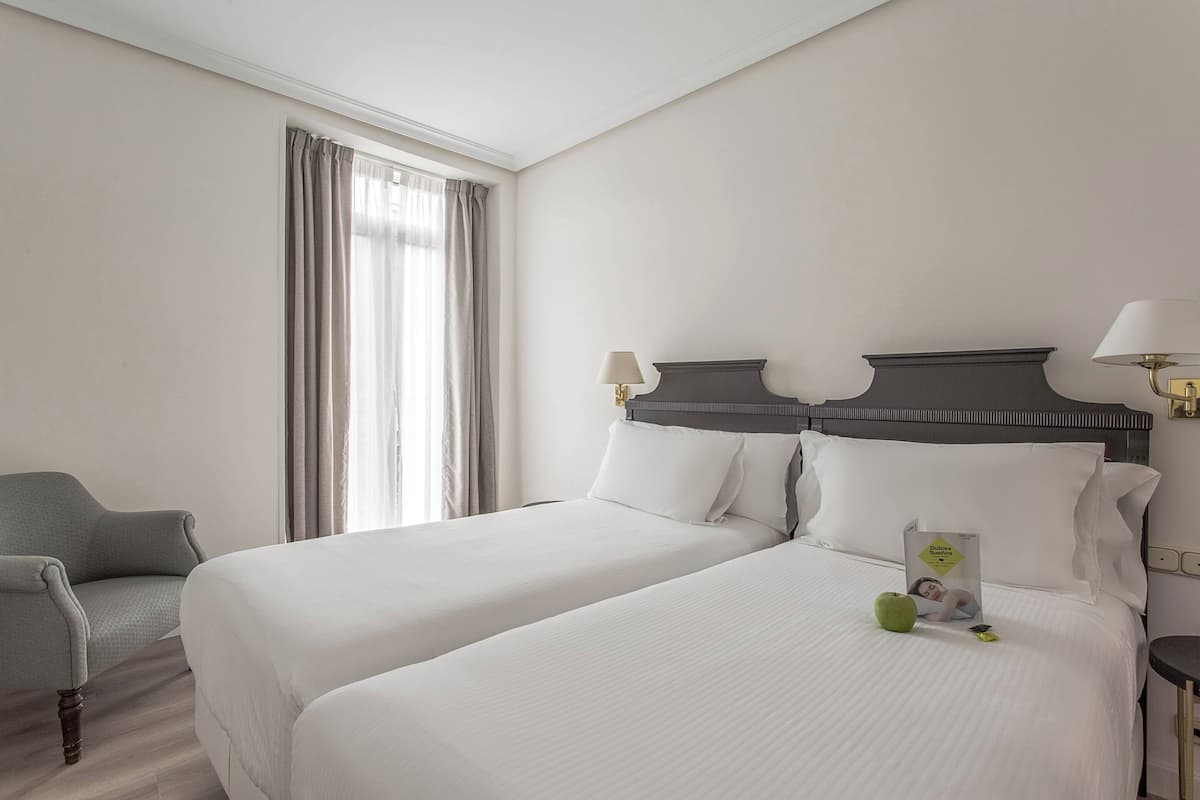 A hotel room with two beds, a chair, a nightstand, and a window with curtains.