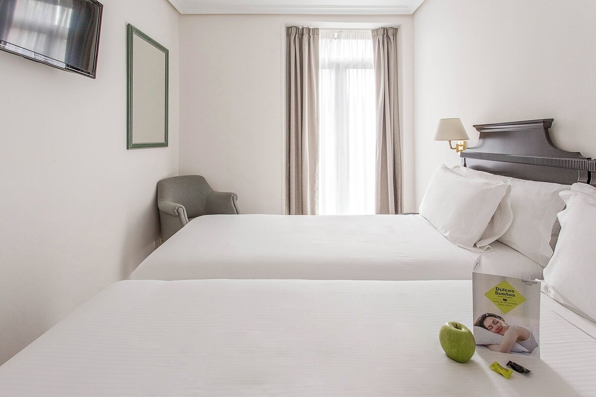 A hotel room with two beds, a TV, a chair, and a window with curtains.