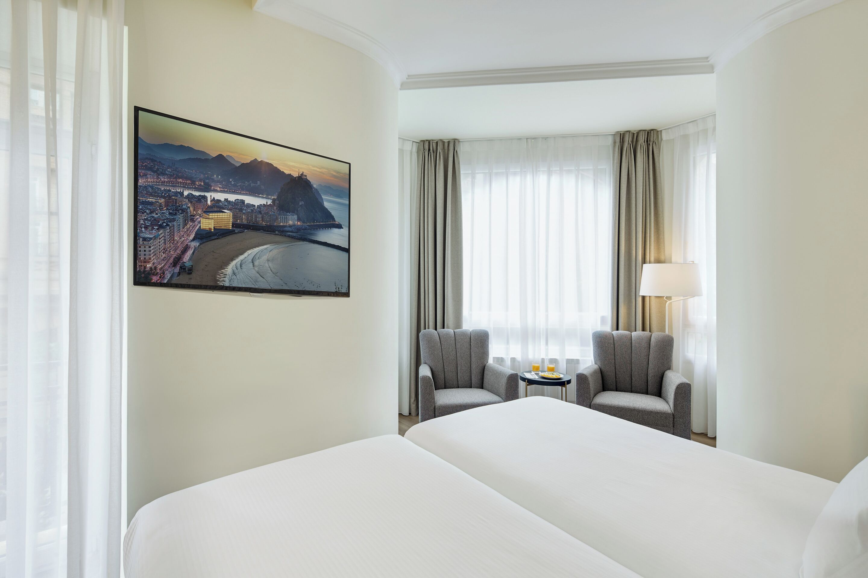 Ein Hotelzimmer mit einem großen Bett, zwei Sesseln, einem kleinen Tisch mit Lampe und einem Stadtbild an der Wand.