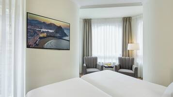 Ein Hotelzimmer mit einem großen Bett, zwei Sesseln, einem kleinen Tisch mit Lampe und einem Stadtbild an der Wand.
