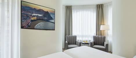 A hotel room with a large bed, two armchairs, a small table with a lamp, and a cityscape painting on the wall.