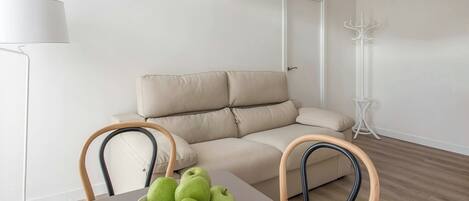 Una sala de estar minimalista con un sofá color beige, una mesa blanca con un tazón de manzanas verdes y una lámpara de pie.