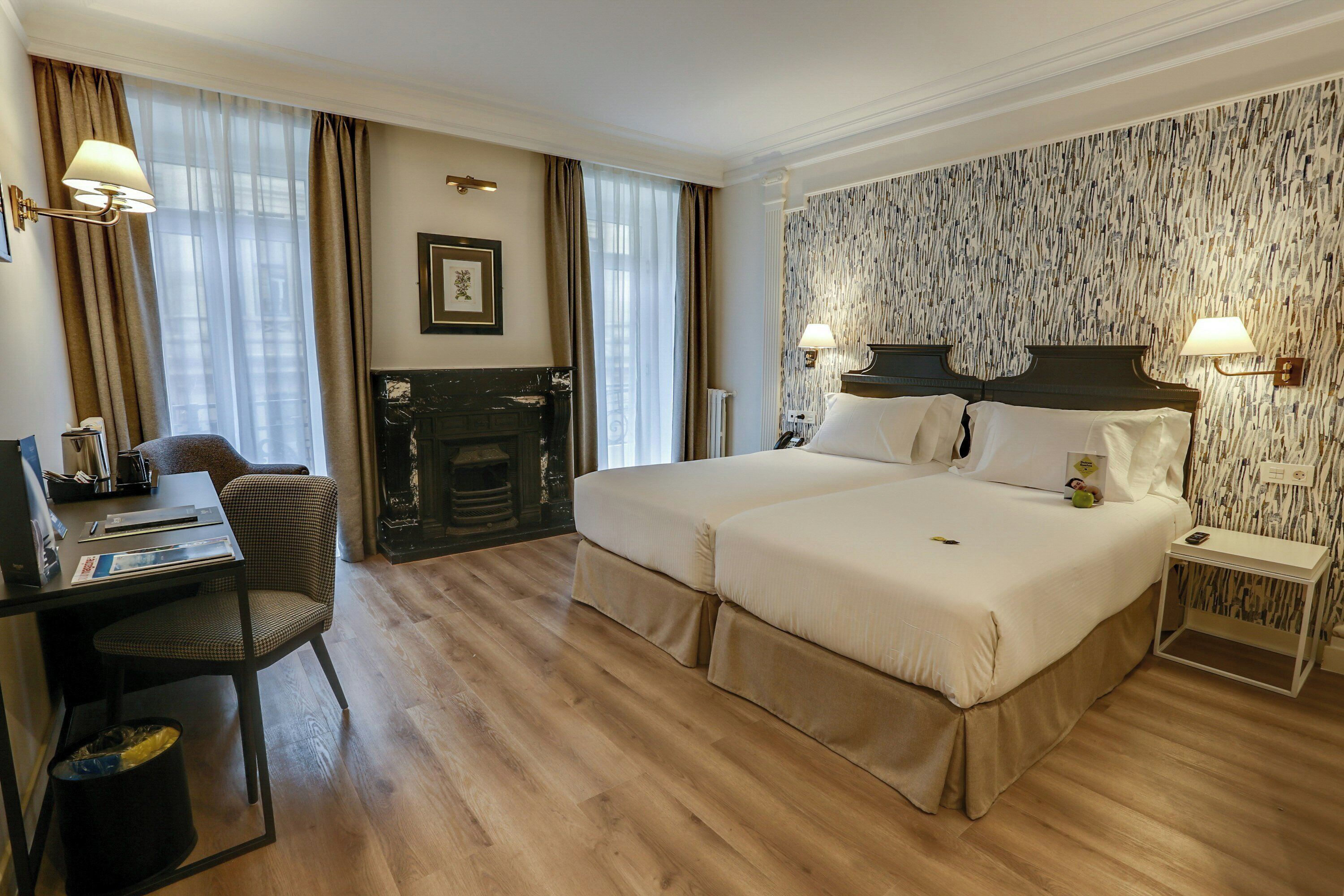 A hotel room with a double bed, a desk with a chair, a fireplace, and a patterned wallpaper.