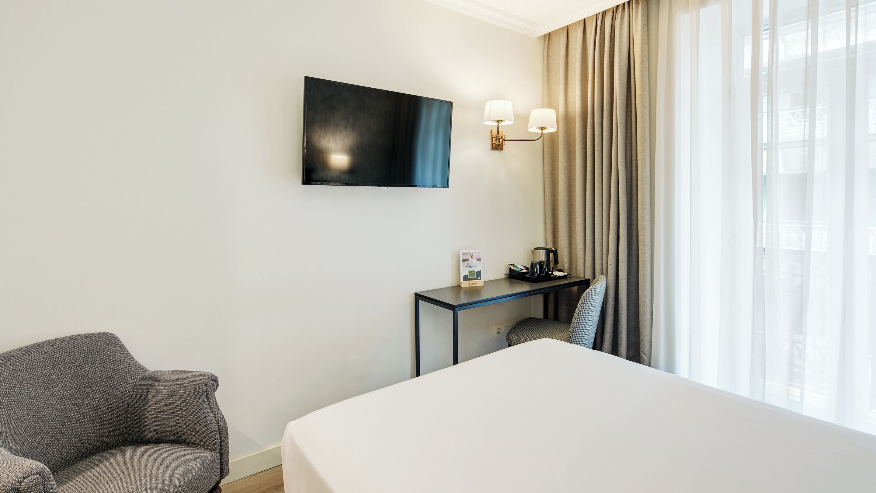 Een hotelkamer met een bed, een bureau met een lamp, een stoel en een televisie die aan de muur is bevestigd.