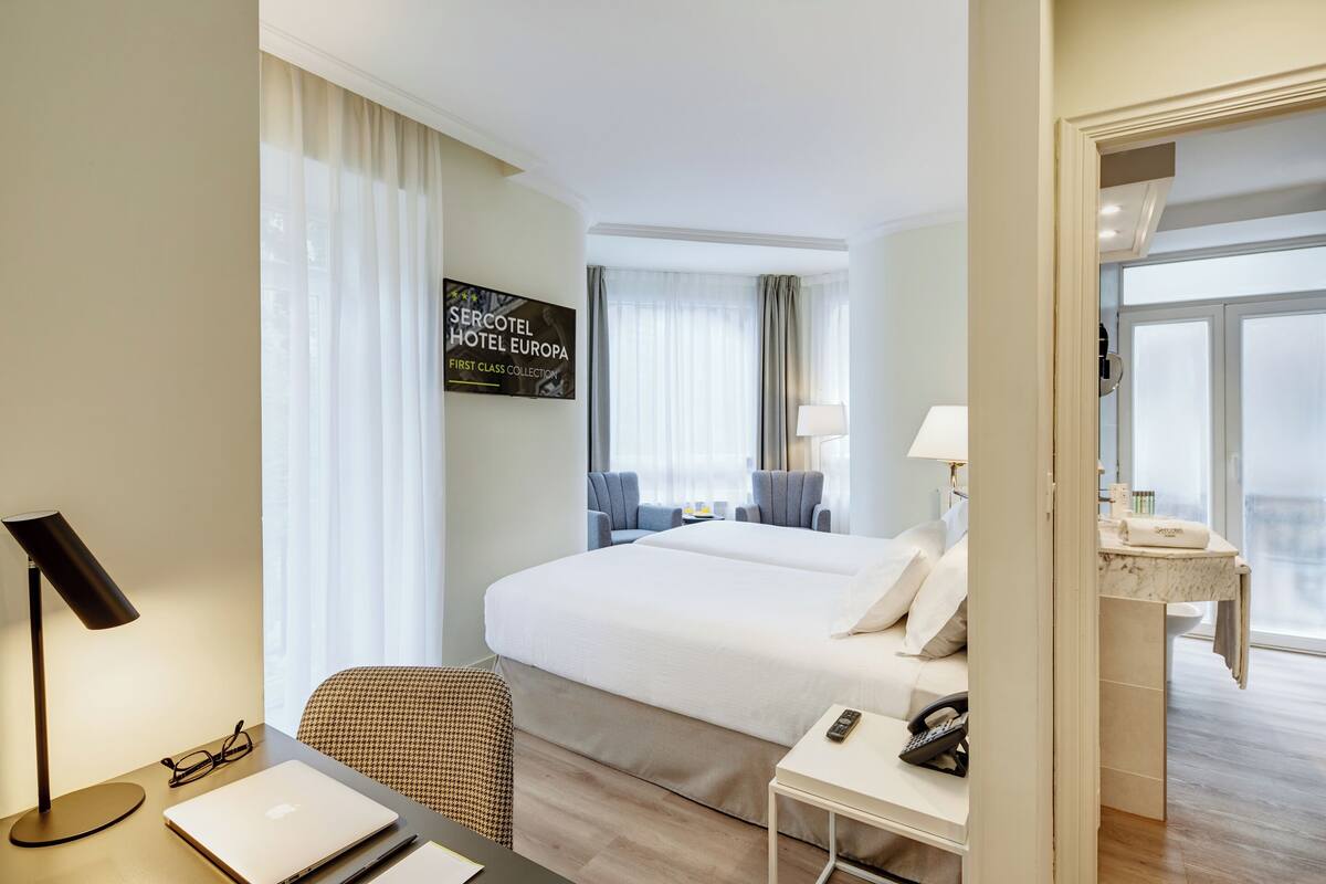 A hotel room with a large bed, a desk with a laptop, a chair, a bedside table with a lamp, and a view of the city through the window.