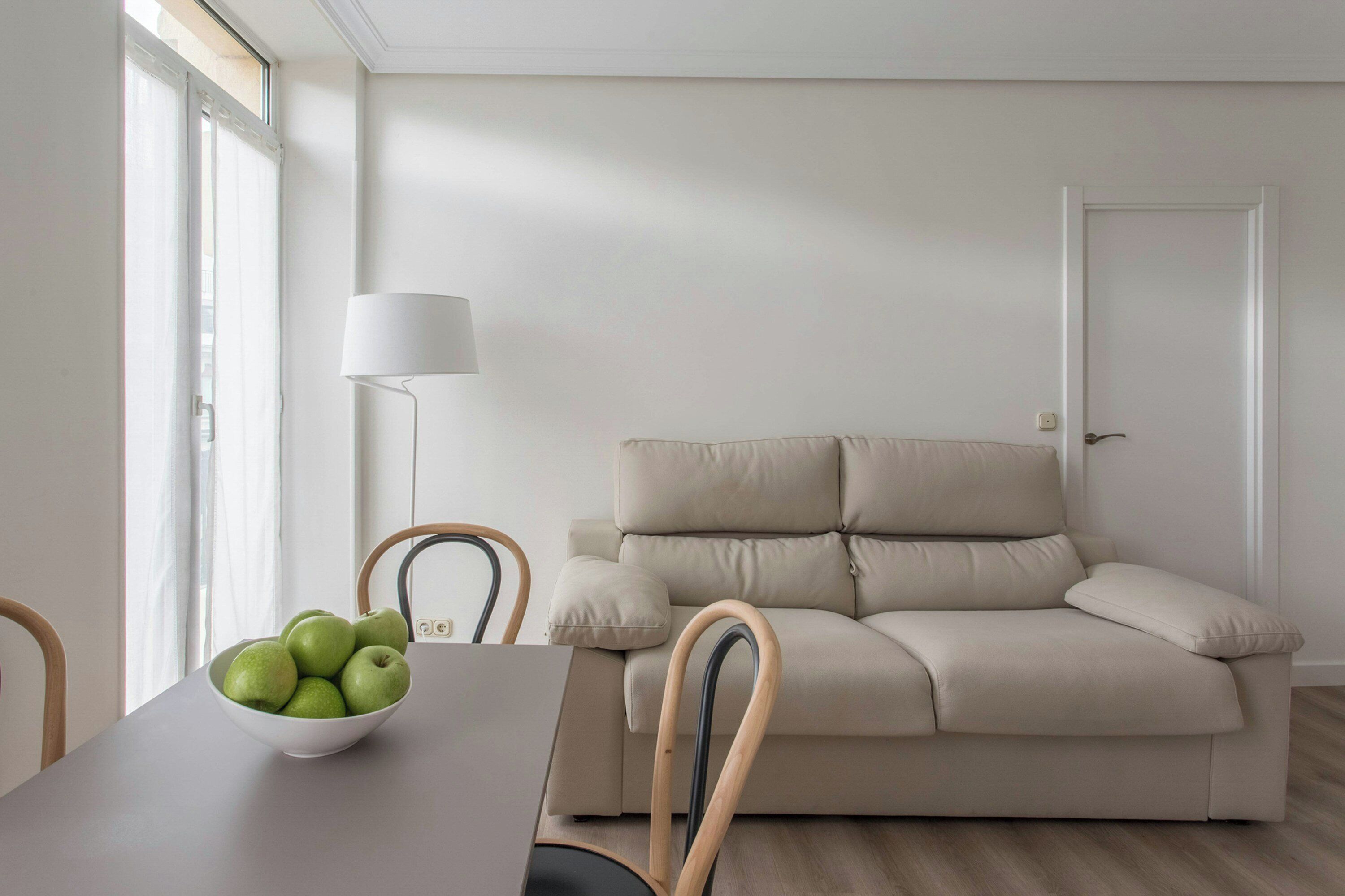 A minimalist living room with a beige sofa, a white table with a bowl of green apples, and a floor lamp.