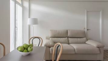 Una sala de estar minimalista con un sofá beige, una mesa blanca con un bol de manzanas verdes y una lámpara de pie.