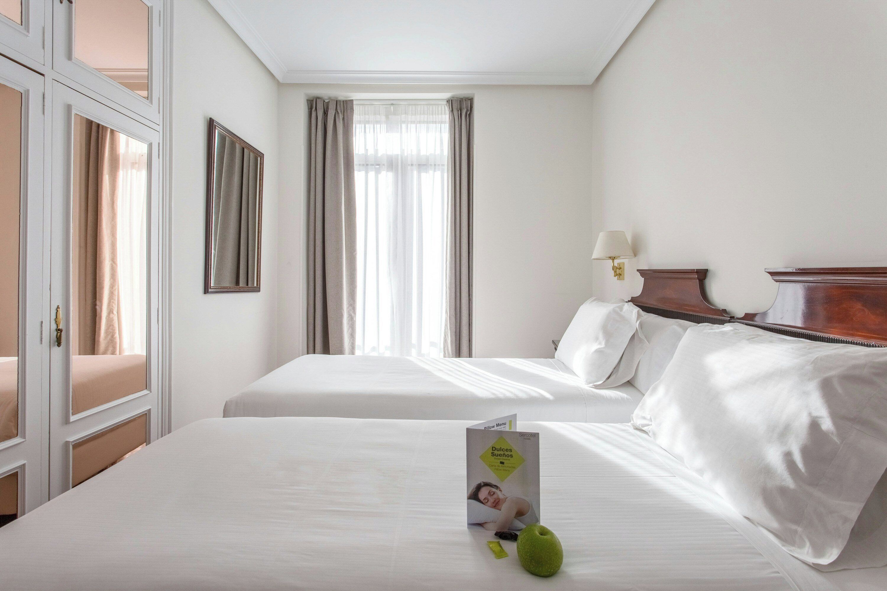 Habitación de hotel con dos camas, una ventana con cortinas, una lámpara de noche y un menú de almohadas sobre la cama.