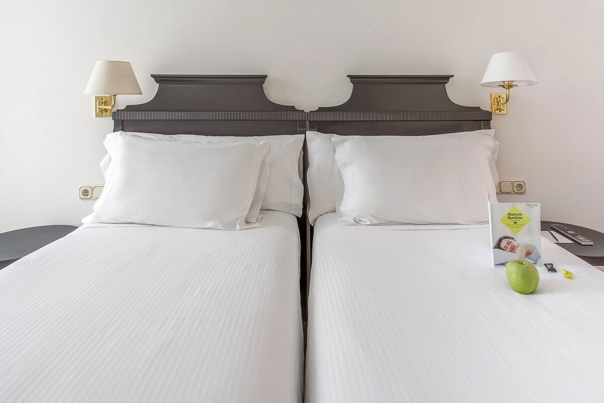 Una cama doble con sábanas blancas, dos lámparas de noche, una mesita de noche con un control remoto y una manzana verde sobre la mesita.