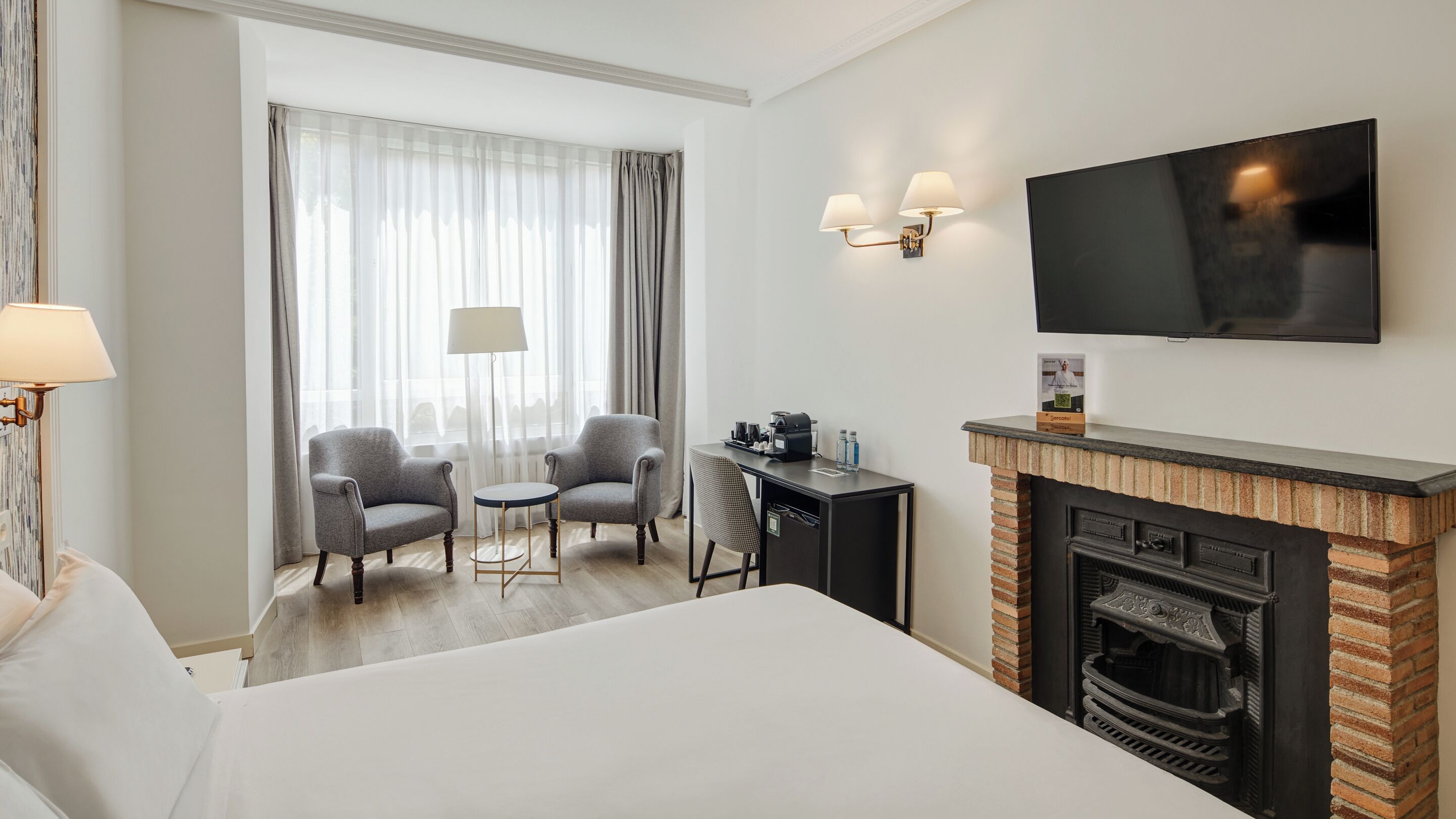 Une chambre d’hôtel comprenant un lit, deux fauteuils, une petite table, une cheminée, un téléviseur et une fenêtre avec des rideaux.