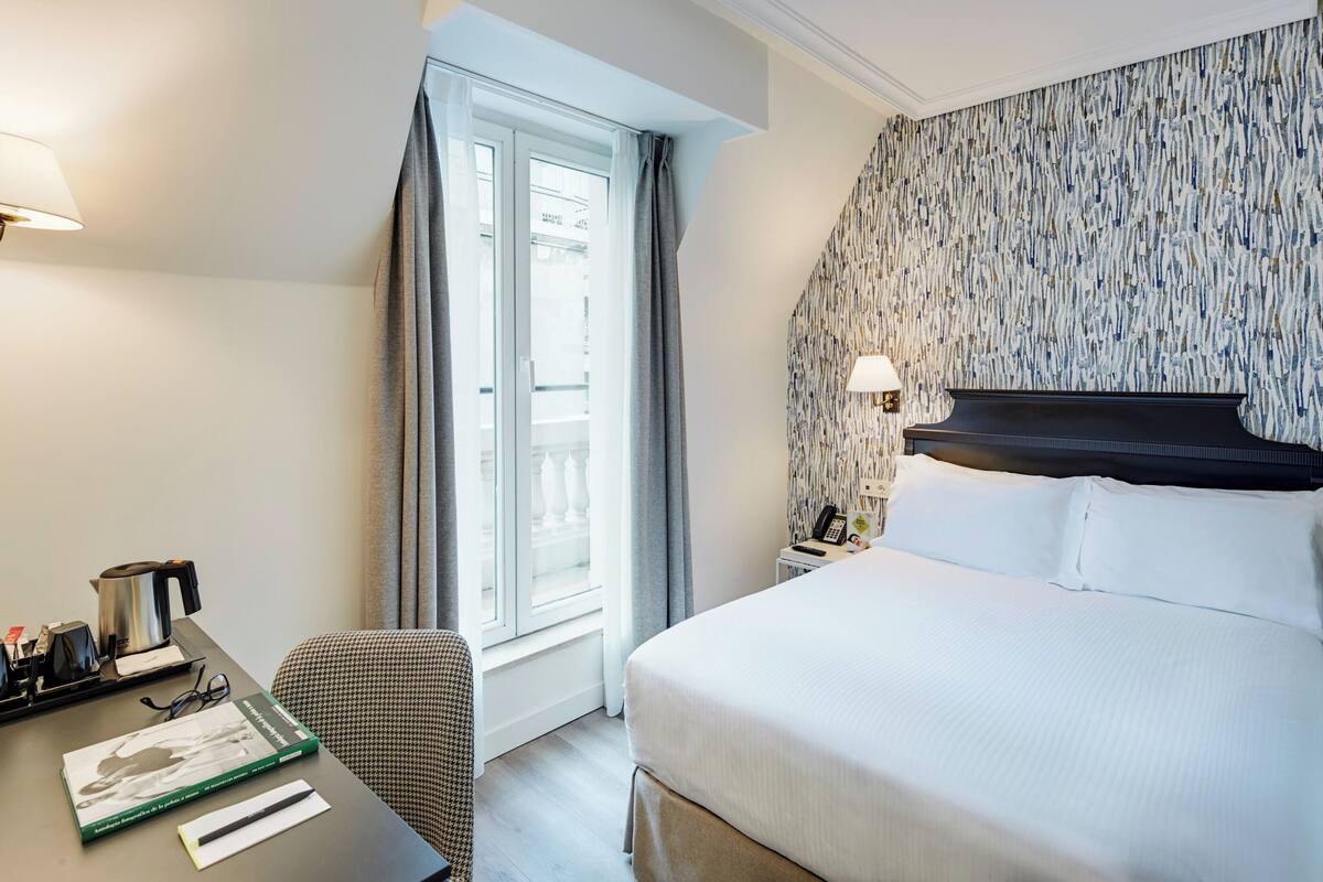 A hotel room with a bed, a desk with a kettle, a chair, a window with curtains, and a patterned wall.