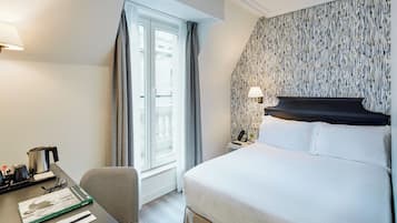 A hotel room with a bed, a desk with a kettle, a chair, a window with curtains, and a patterned wall.