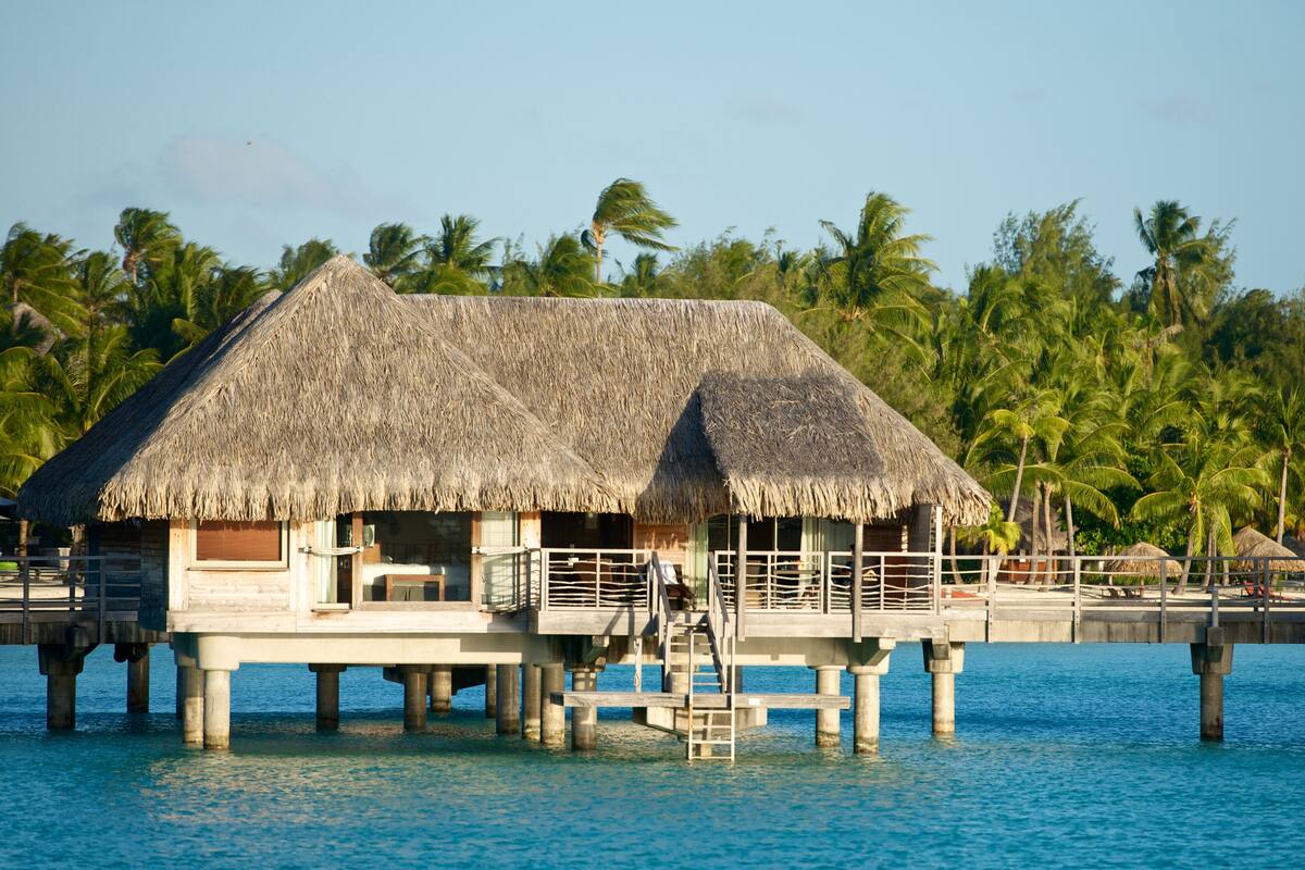 Een overwaterbungalow met rieten dak, houten terras en een trap naar het bovenste niveau.