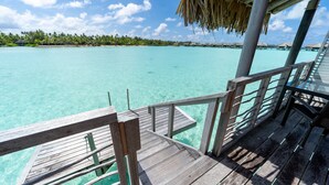 Een houten terras met een balustrade dat uitkijkt over turquoise water, een rieten dakconstructie en een verre kustlijn met waterwoningen.