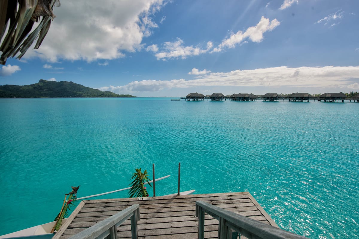Een houten terras met uitzicht op turkoois water, met drijvende bungalows in de verte.