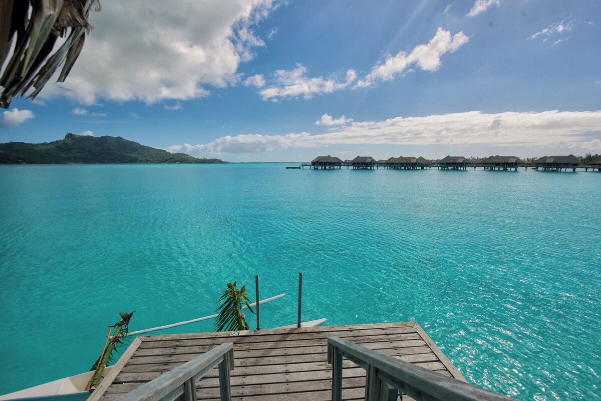 Terraza de madera con vistas a aguas turquesas y bungalows sobre el agua en la distancia.