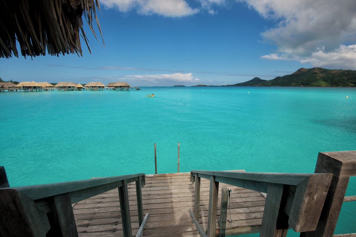 Een houten terras dat uitkomt op turquoise water, met drijvende bungalows op de achtergrond.