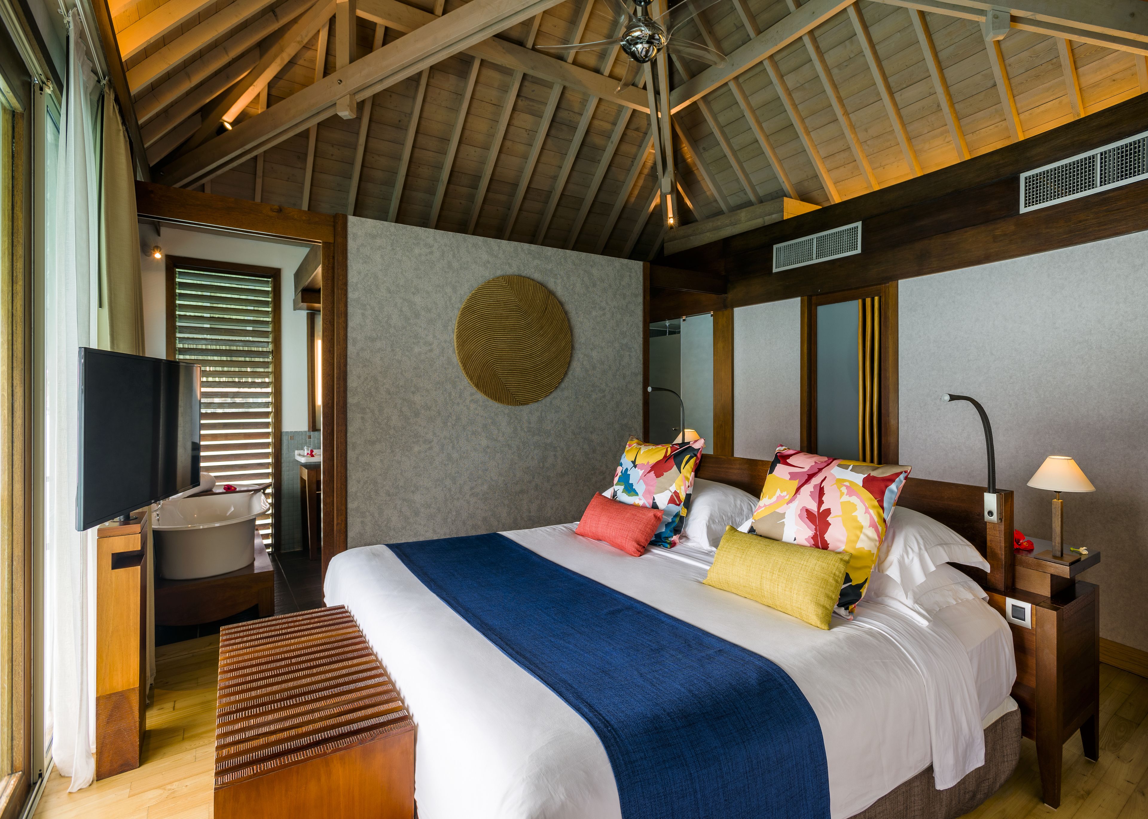 A spacious bedroom with a large bed, a wooden ceiling, a bedside table with a lamp, and a bathroom visible through a window.