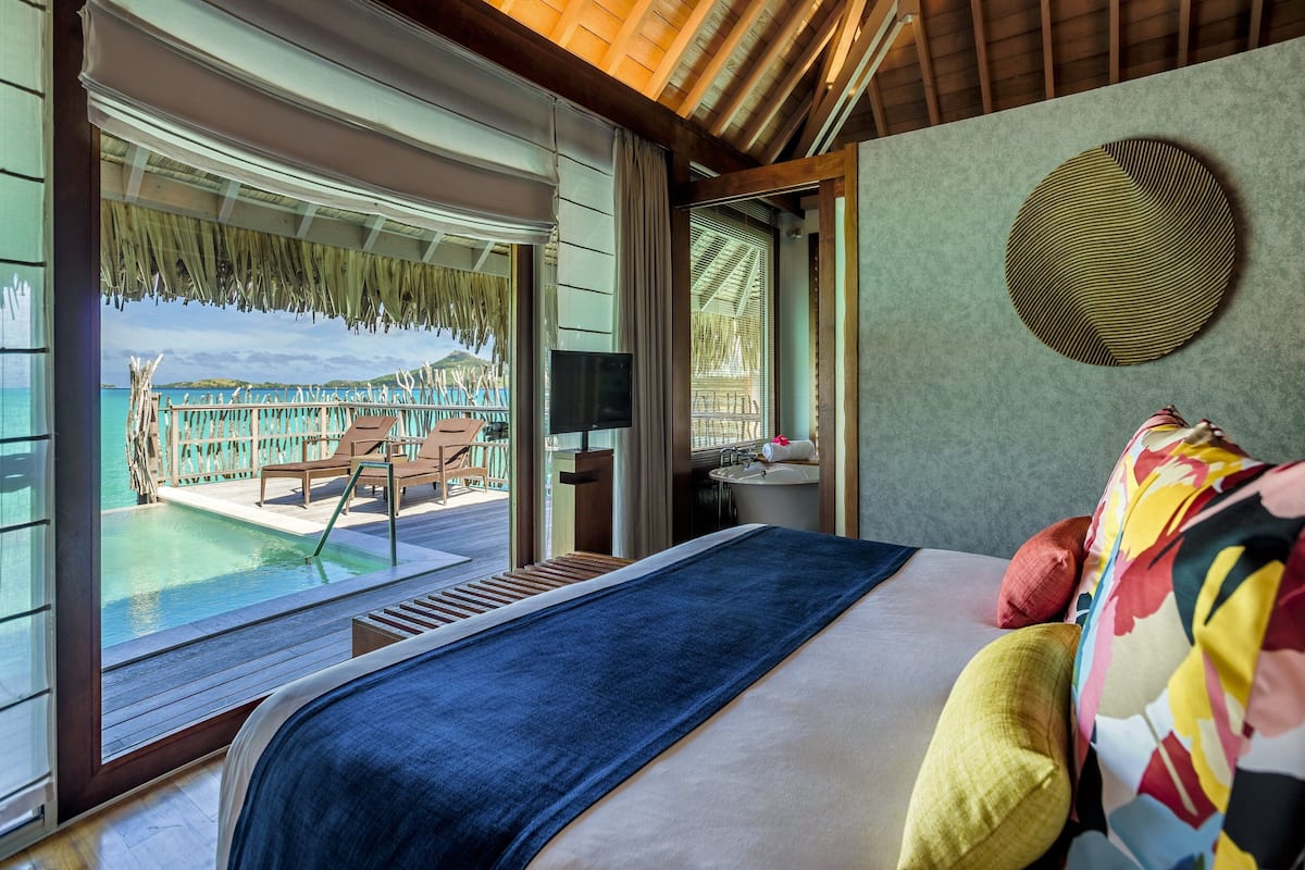 Een slaapkamer met een groot bed, uitzicht op een zwembad en strand, en een badkamer met wastafel en spiegel.