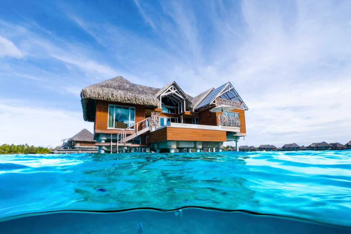 A large, multi-story overwater bungalow with a thatched roof, built on stilts over clear turquoise water.