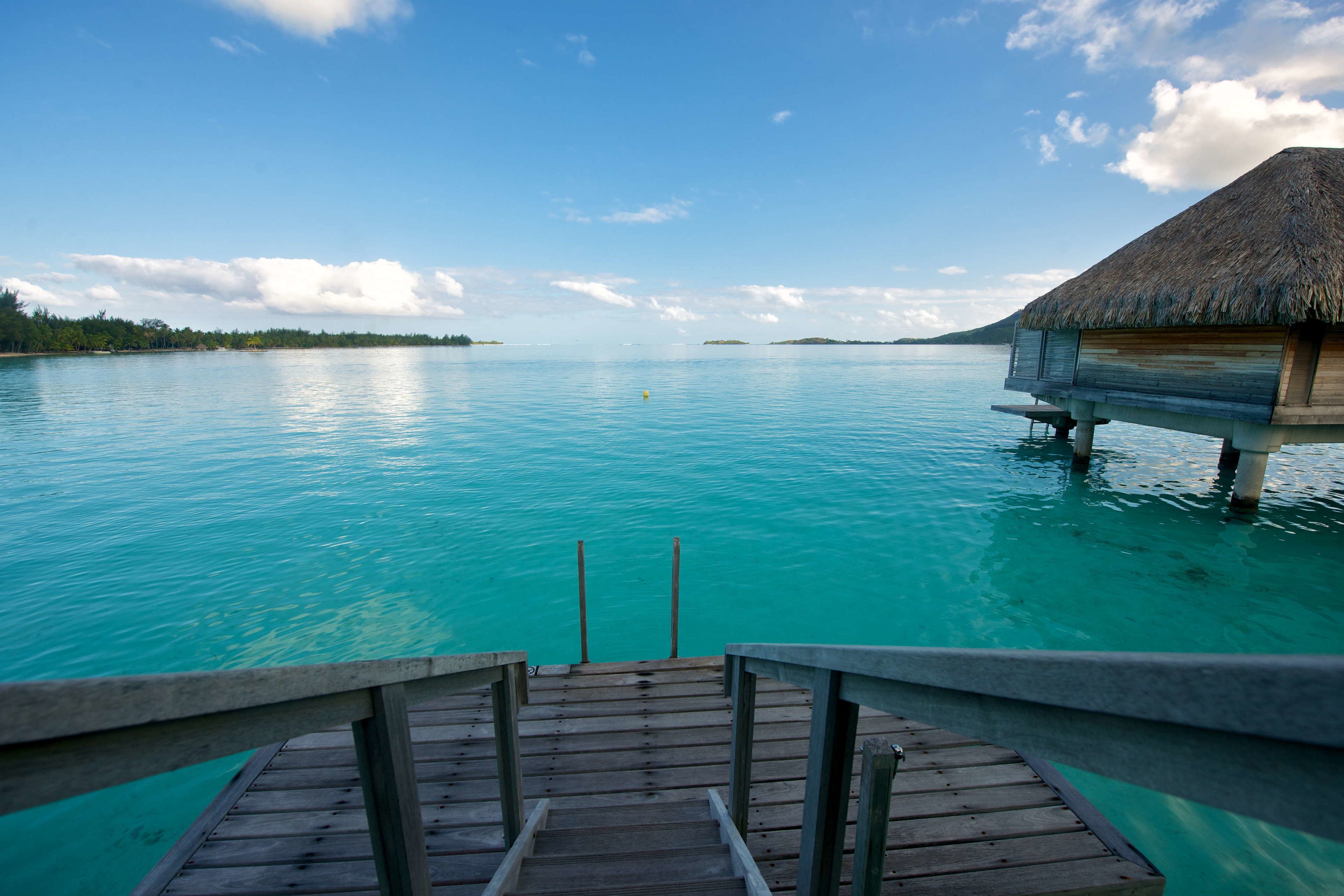 A wooden deck leading to an overwater bungalow with a thatched roof, surrounded by turquoise water and distant islands.
