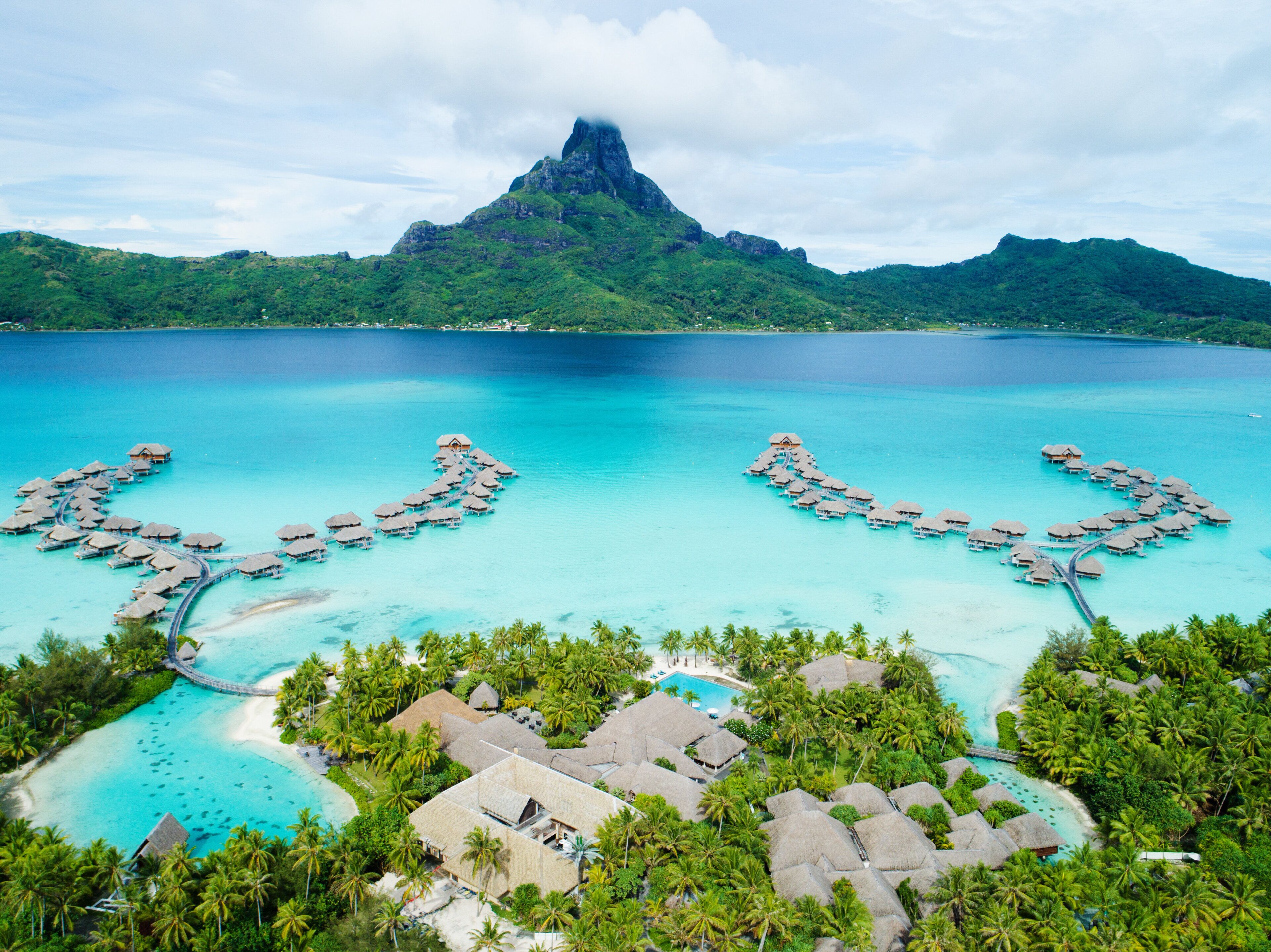 Vista aérea de un resort con bungalows sobre el agua, un mar turquesa cristalino y una montaña imponente al fondo.