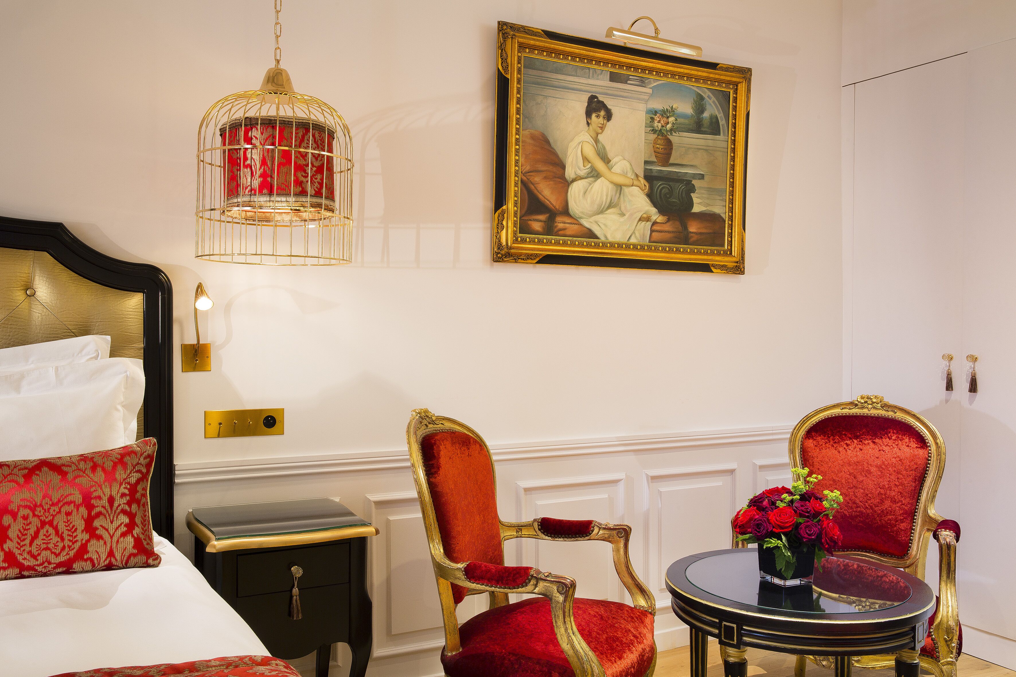 A hotel room with a bed, two red upholstered chairs, a small table with a vase of red roses, a gold-framed painting, and a hanging light fixture.