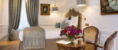 A hotel room with a bed, a chair, a table with a vase of flowers, a picture on the wall, and a window with curtains.