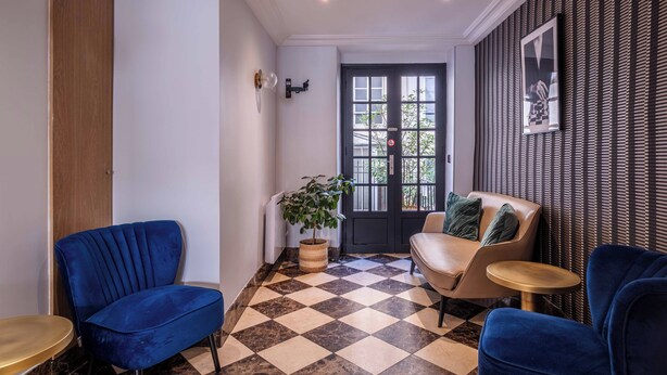 Een kamer met een geruit vloerkleed, twee blauwe fauteuils, een beige bank, een klein rond tafeltje en een potplant.
