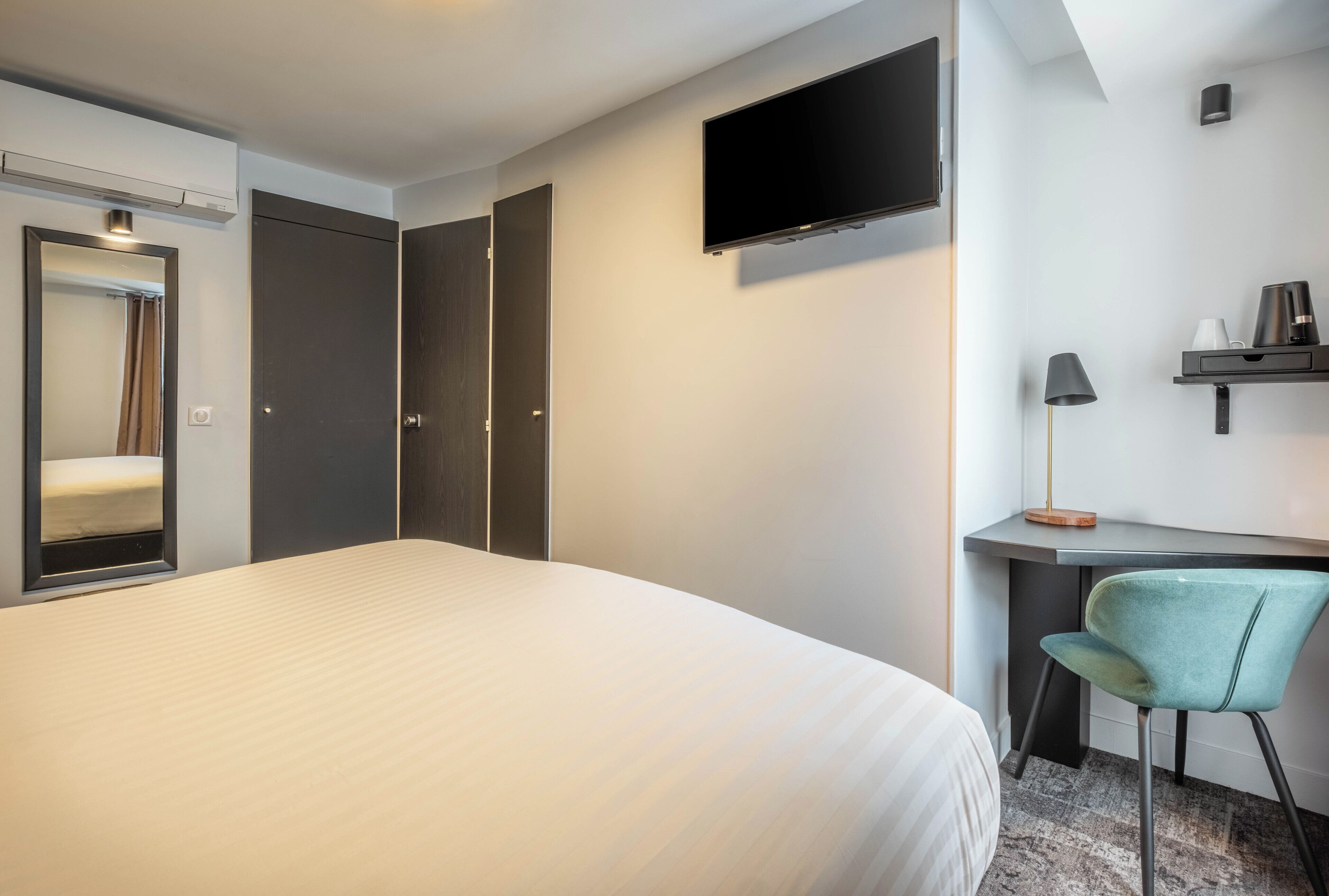 A hotel room with a large bed, a desk with a chair, a TV mounted on the wall, and a wardrobe.
