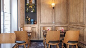 Een eethoek met houten stoelen en tafels, een wandplank met decoratieve voorwerpen, en een deur met geëtste glaspanelen.