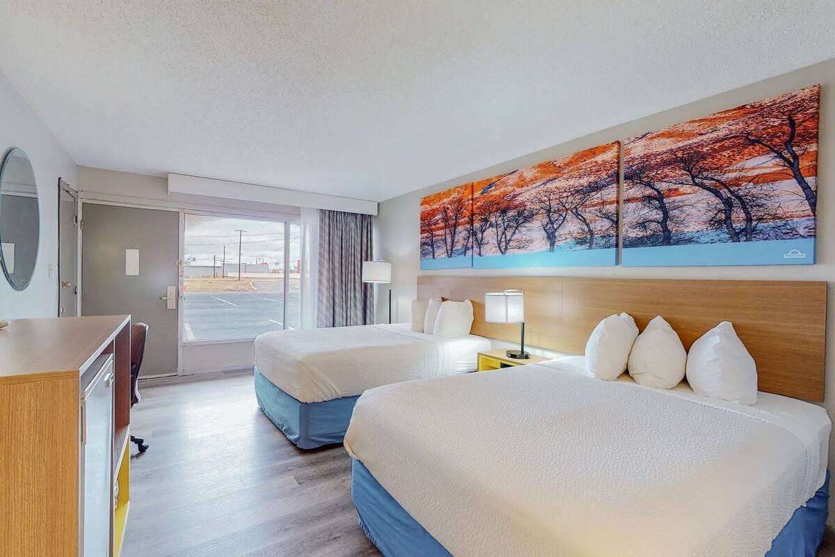 Quarto de hotel com duas camas, uma escrivaninha, um espelho, um quadro e vista para o estacionamento.