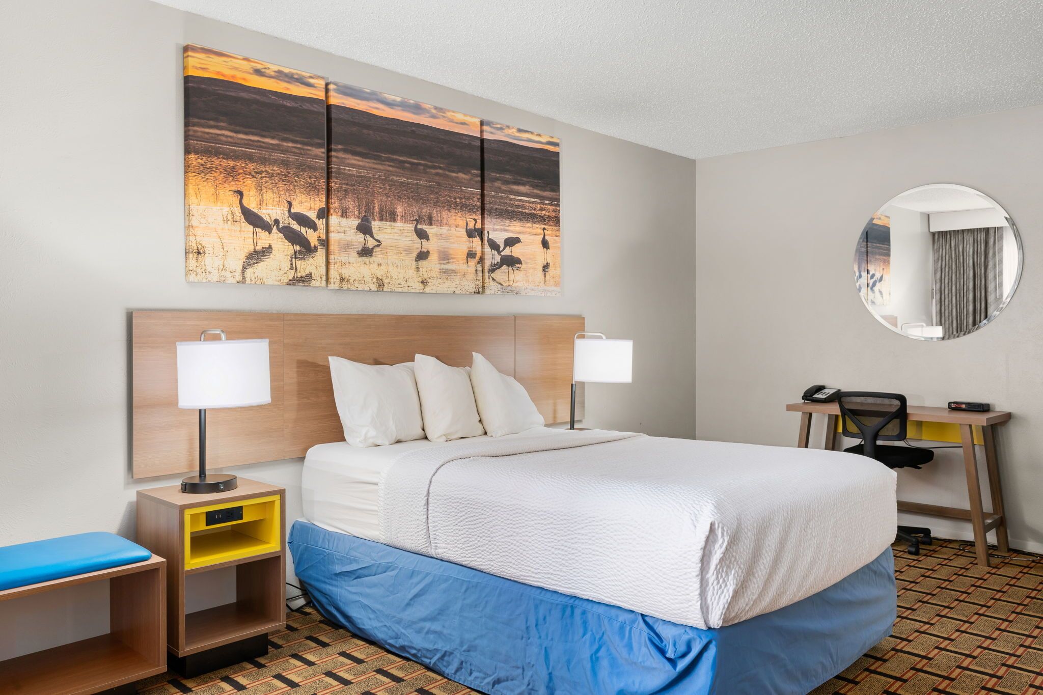 A hotel room with a large bed, two bedside tables, a desk with a chair, a round mirror, and a wall-mounted artwork of birds.
