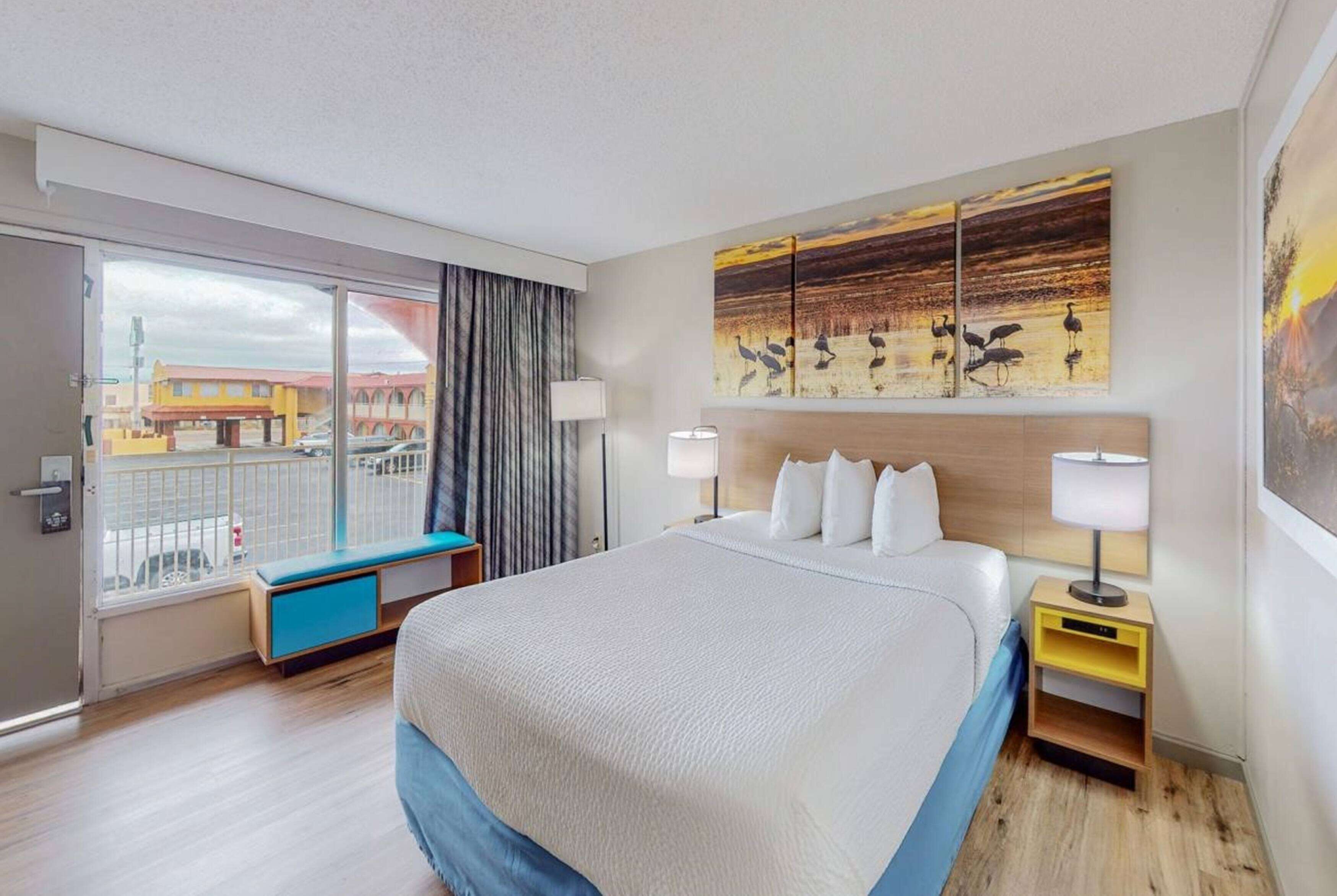 A hotel room with a large bed, a view of the city through a sliding door, a bench by the window, and artwork on the walls.