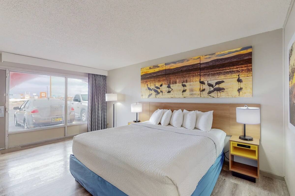 Quarto de hotel com uma cama grande, duas mesas de cabeceira com abajures, vista da cidade através de uma porta de correr e um quadro de pássaros na parede.