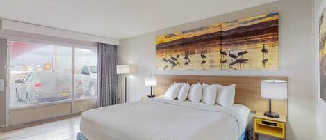 A hotel room with a large bed, two bedside tables with lamps, a view of the city through a sliding door, and a painting of birds on the wall.