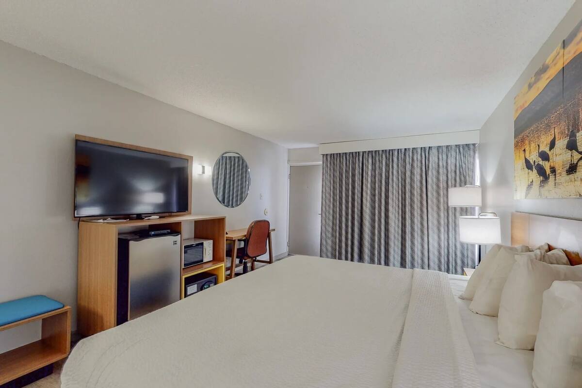 Quarto de hotel com cama, televisão, escrivaninha, cadeira, abajur e um quadro na parede.