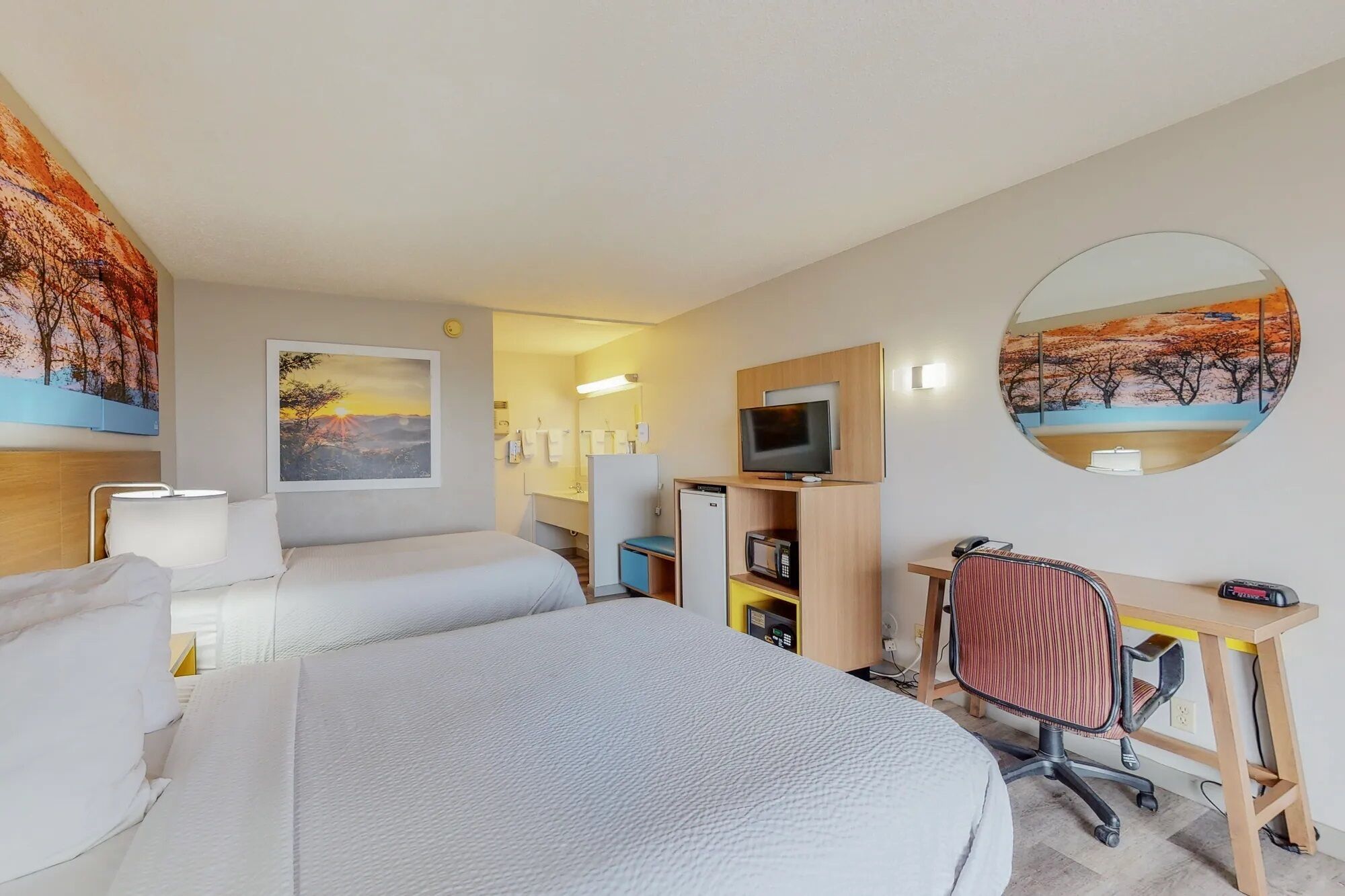 Habitación de hotel con cama, escritorio, silla, televisor, microondas y baño a la vista a través de una puerta abierta.